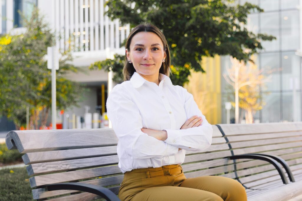 Woman sitting confidently on a bench in an urban park setting, wearing a white shirt.